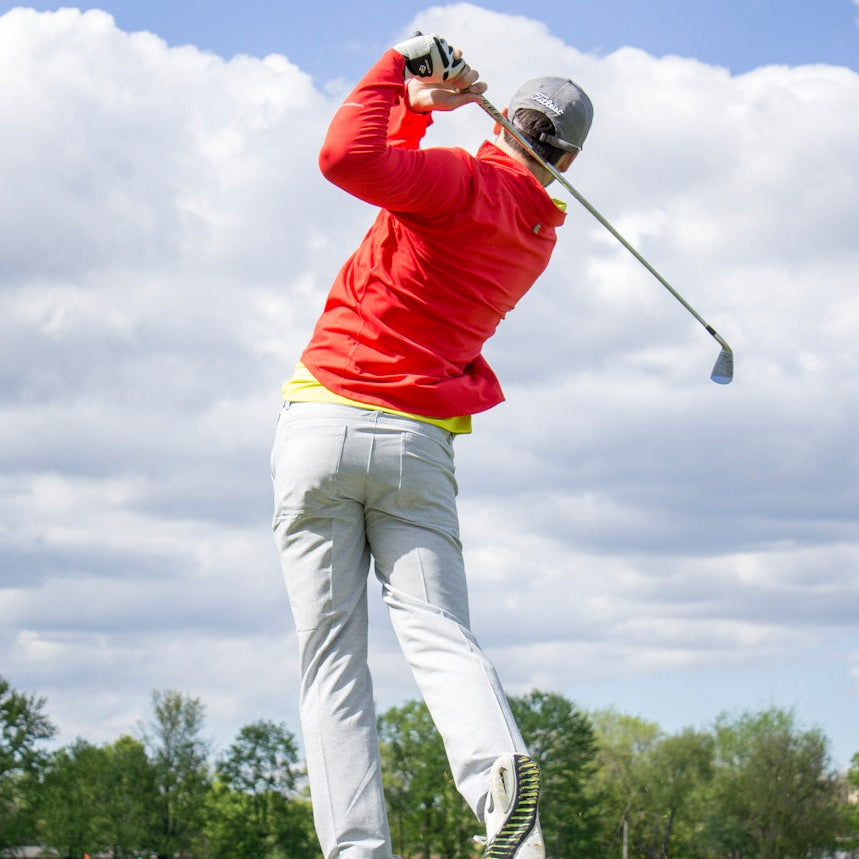Person playing golf with a red jacket and white pants against a cloudy sky.