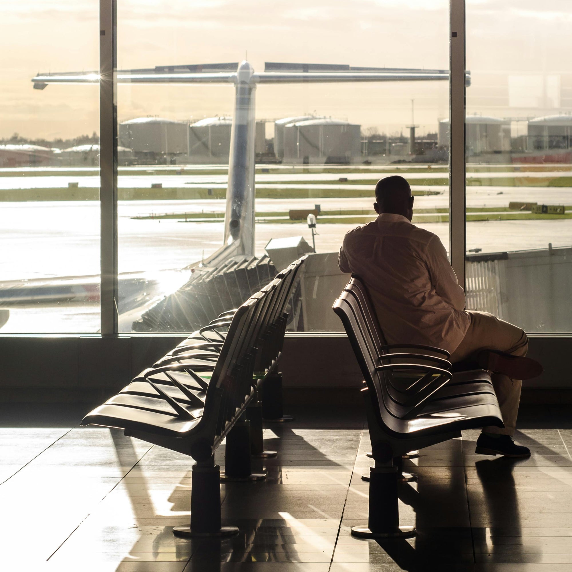 Person sitting on a bench in an airport terminal looking at an airplane through large windows.